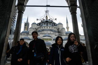 People in dark clothes standing in the foreground of a grand white mosque with blue domed roofs