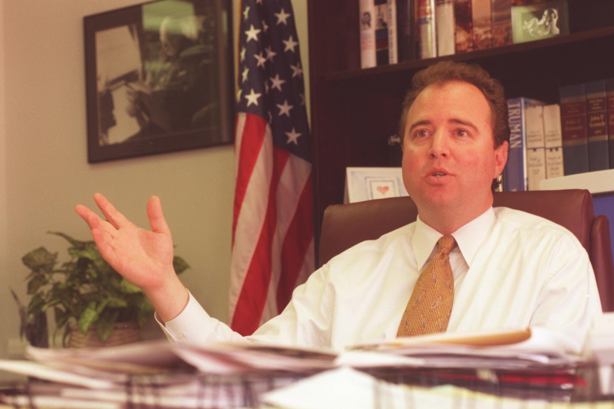 Adam Schiff gestures as he speaks from behind a desk piled with paperwork, a bookcase and American flag behind him