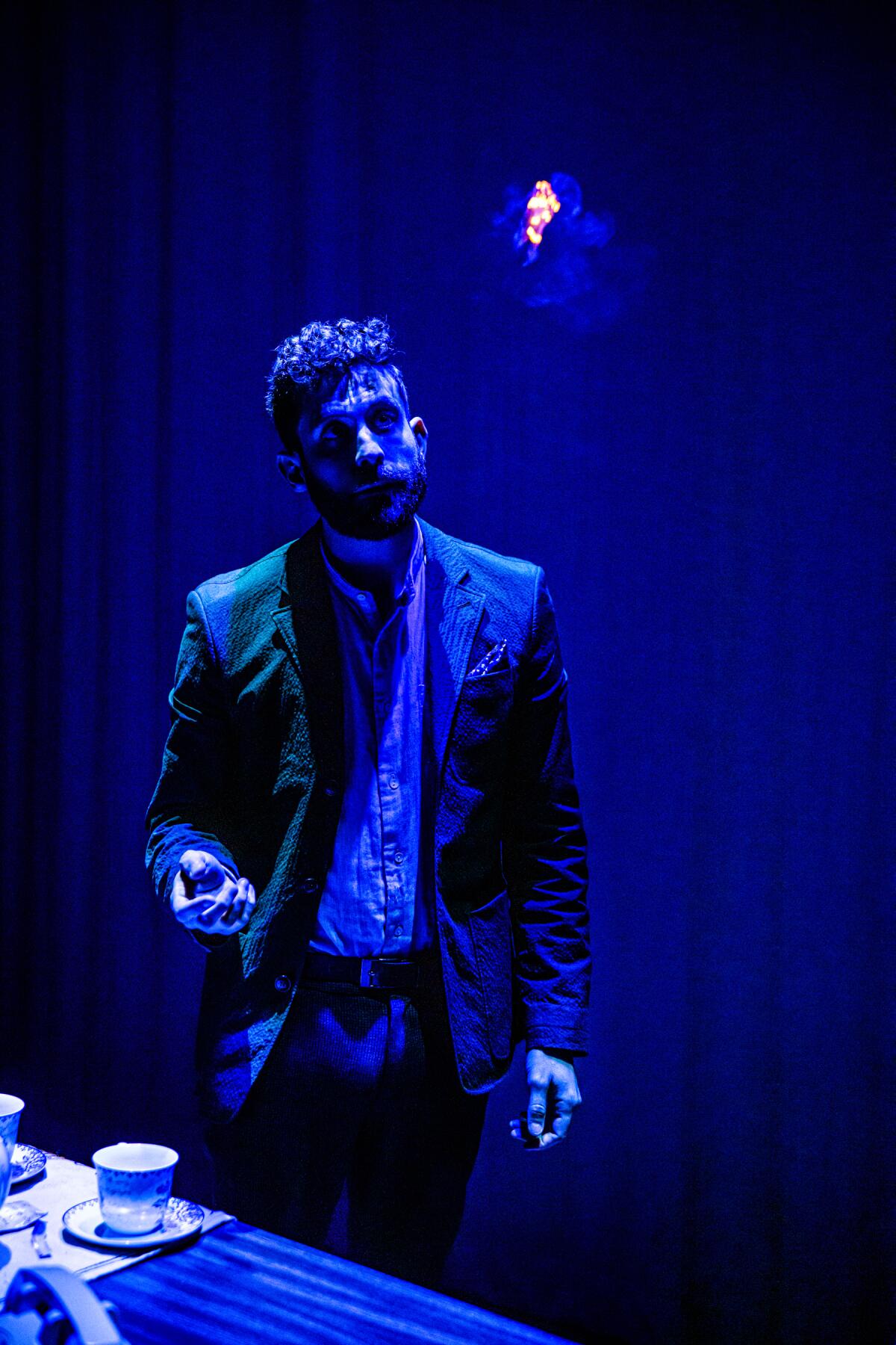 A man in dark lighting gestures near a table 