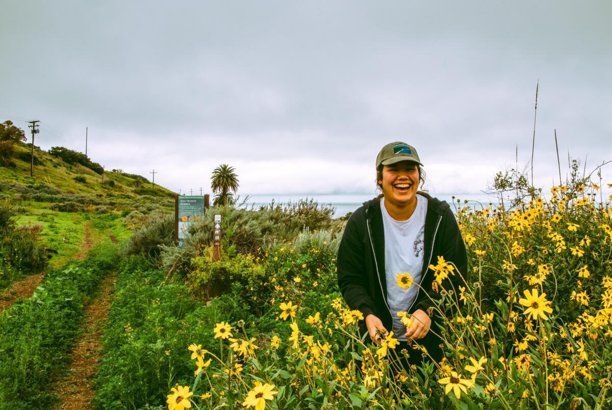 A woman in a baseball cap stands among yellow flowering plants with the ocean behind her