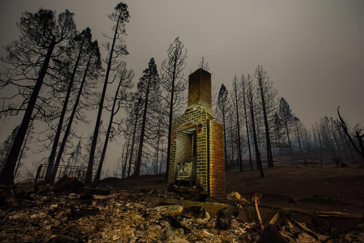 A lone brick chimney stands amid charred ruins of a home under a dark, hazy sky