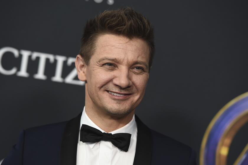 Jeremy Renner smiling in a black suit and bowtie against a black background