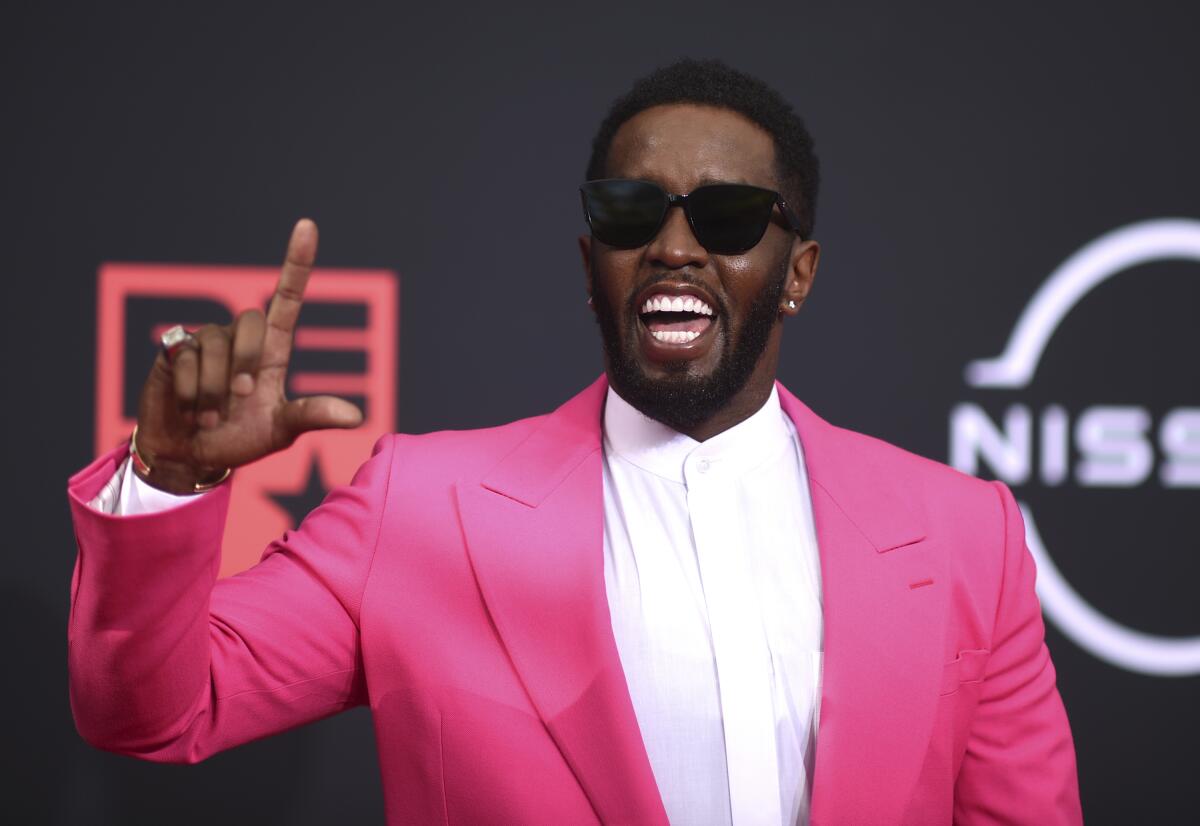 A man wearing a pink suit and sunglasses holds up his index finger as he speaks