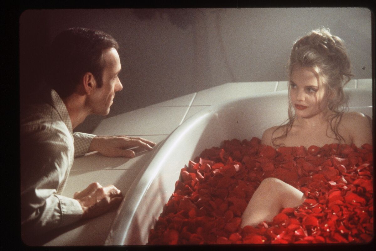 A man faces a woman soaking in a bathtub covered with rose petals.