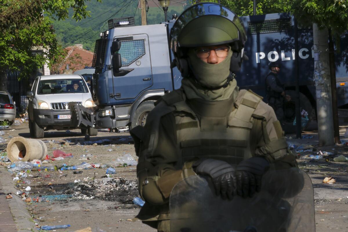 A person wearing glasses, a helmet and olive-colored uniform holds a shield near a police truck on a debris-strewn road 