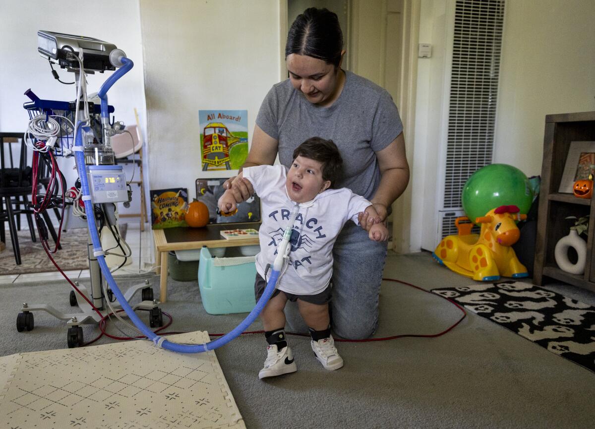 A woman helps a toddler who uses a ventilator walk in their home.