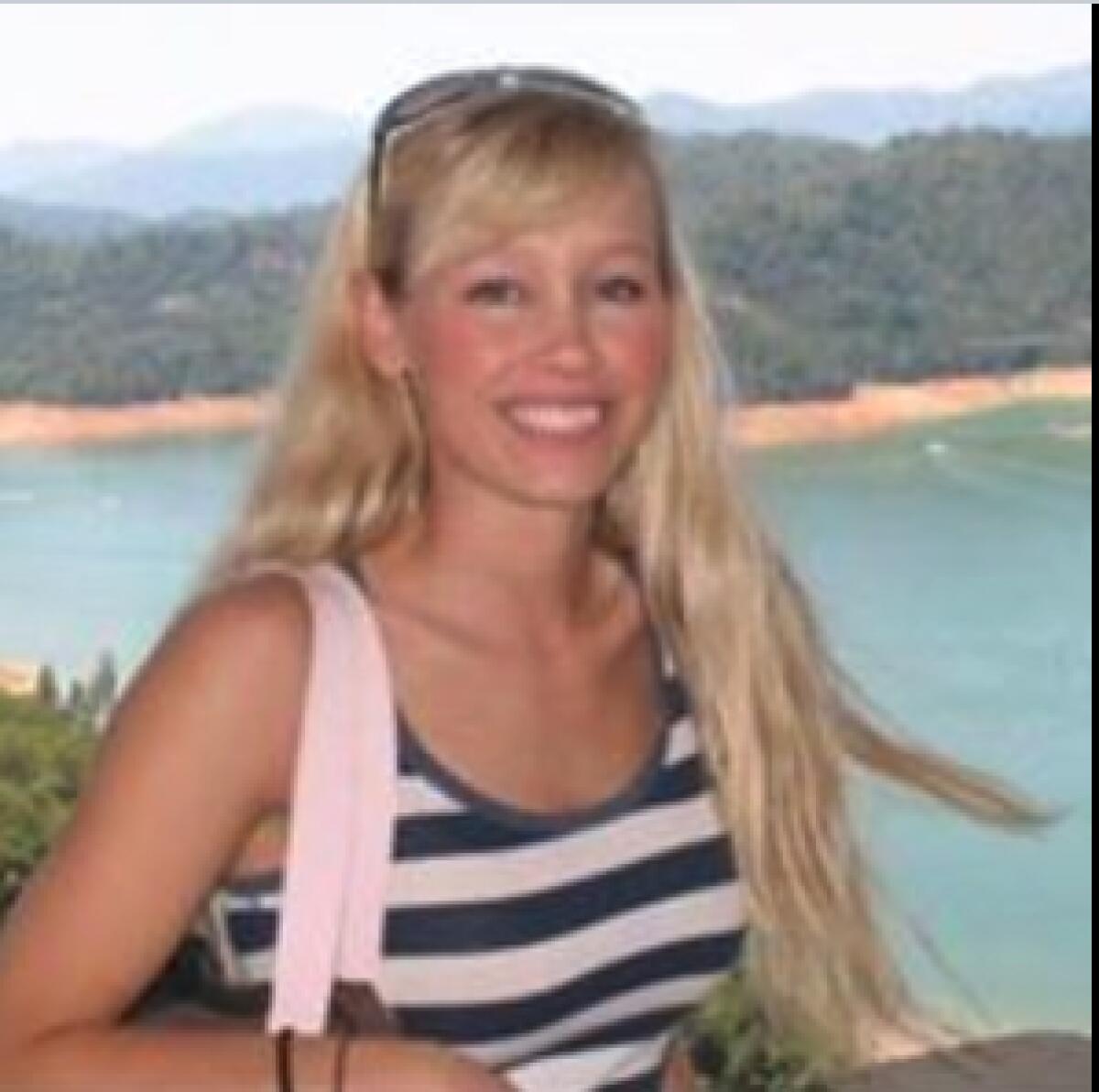 A blond woman smiles for a photo in front of a lake