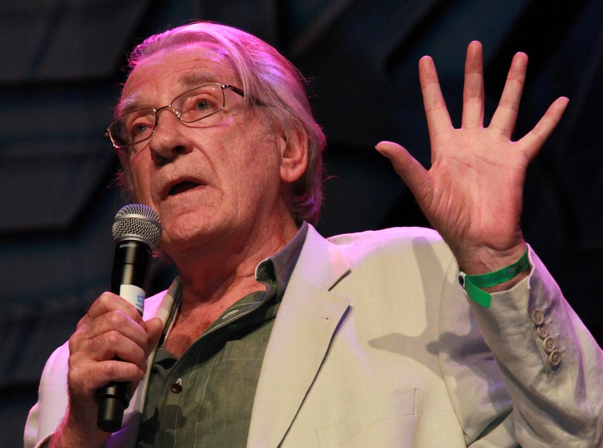 A man wearing glasses holds a microphone in one hand while gesturing with the other