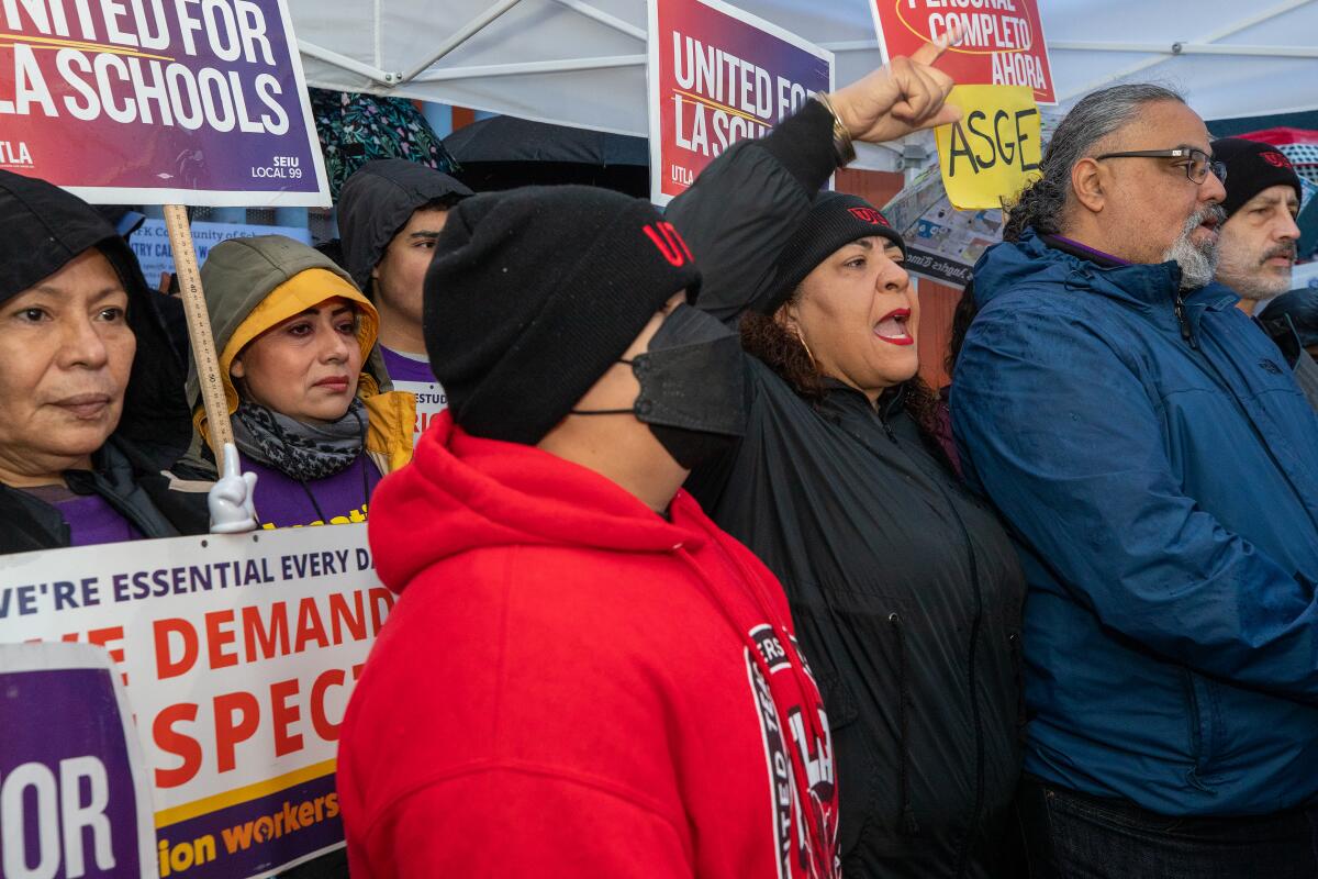 People stand close together. Some hold picket signs. A woman gestures with her hand as she speaks. 