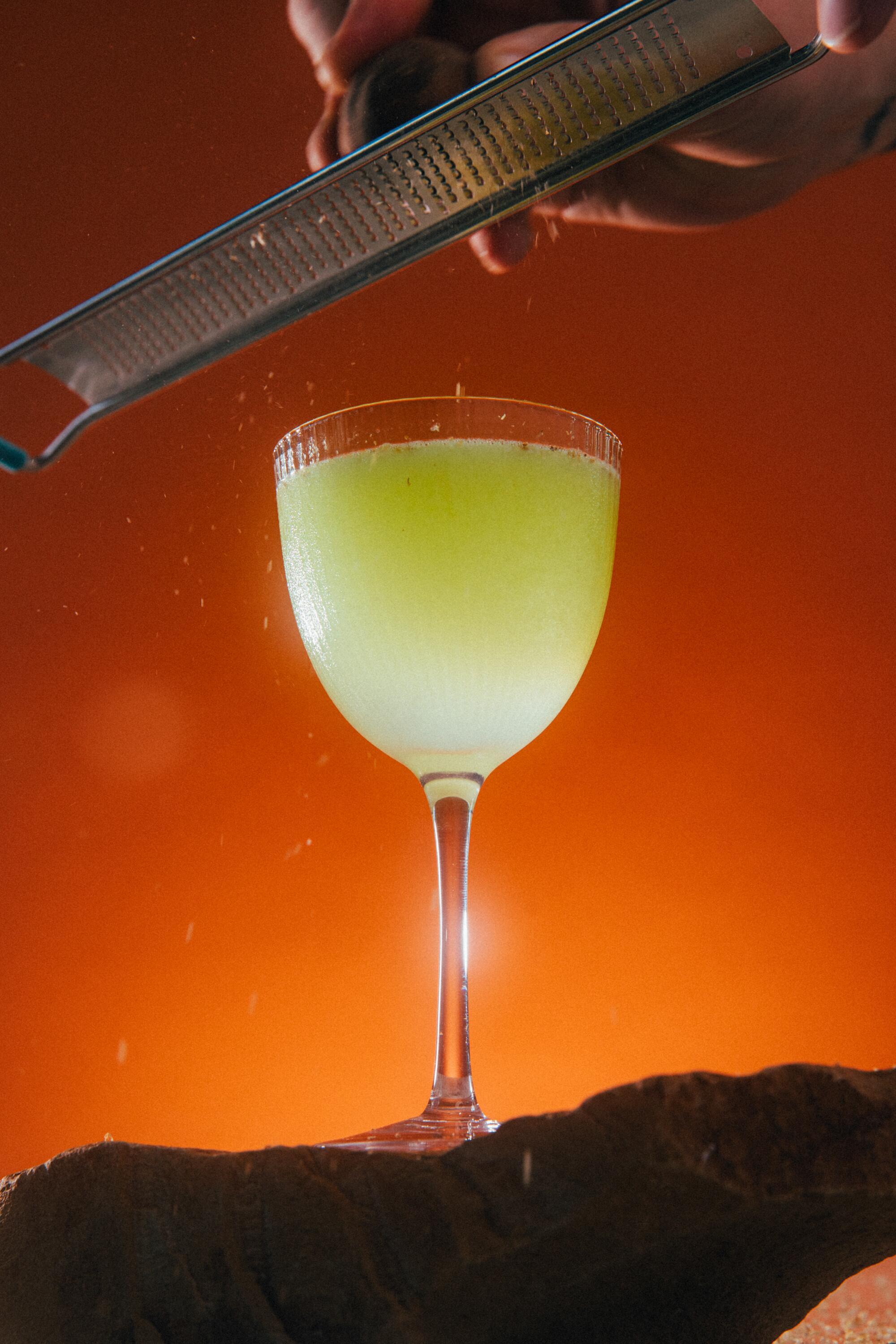 A green cocktail glows in front of an orange background. Above it, a hand grates black-lime zest into the drink.
