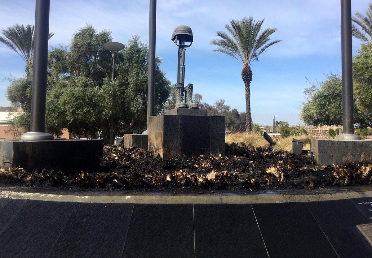 Burned plants surround a statue of boots and a rifle topped by a combat helmet.