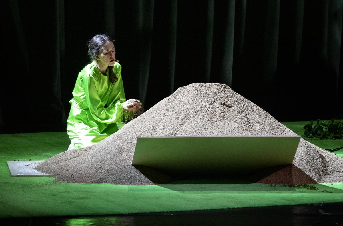 A kneeling woman sifts sand through her hands on a stage bathed in green.