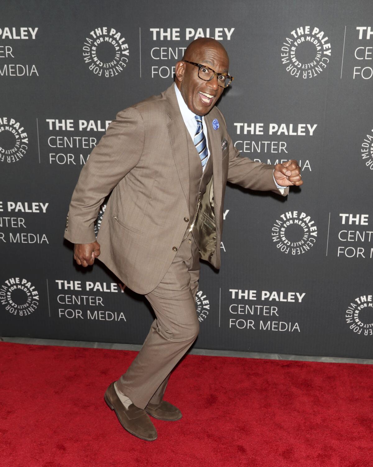 A man in a brown three-piece suit smiles and goofs around on a red carpet