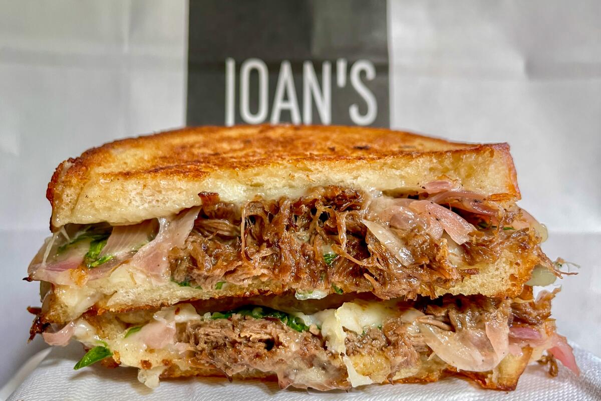 Two stacked halves of a short rib grilled cheese in front of a Joan's on Third sign