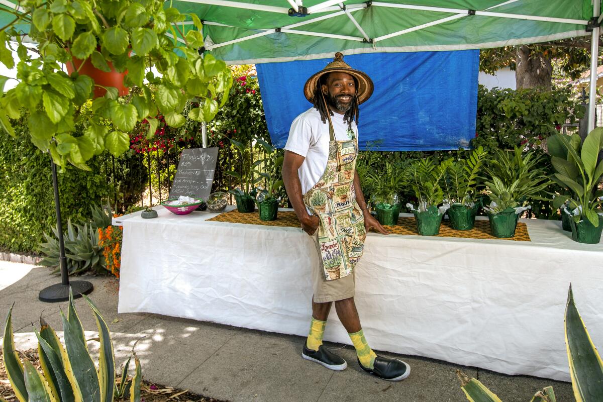 A man in khaki shorts and a plant-patterned apron stands amid houseplants.