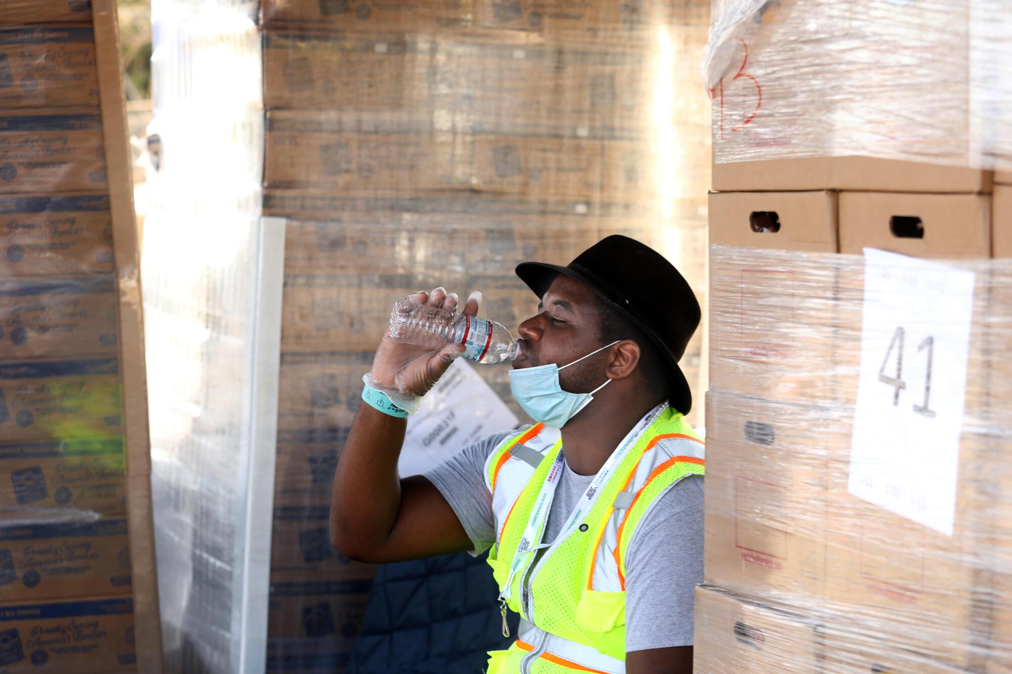 A man in a high-vis vest drinks water while sitting among pallets of cardboard boxes
