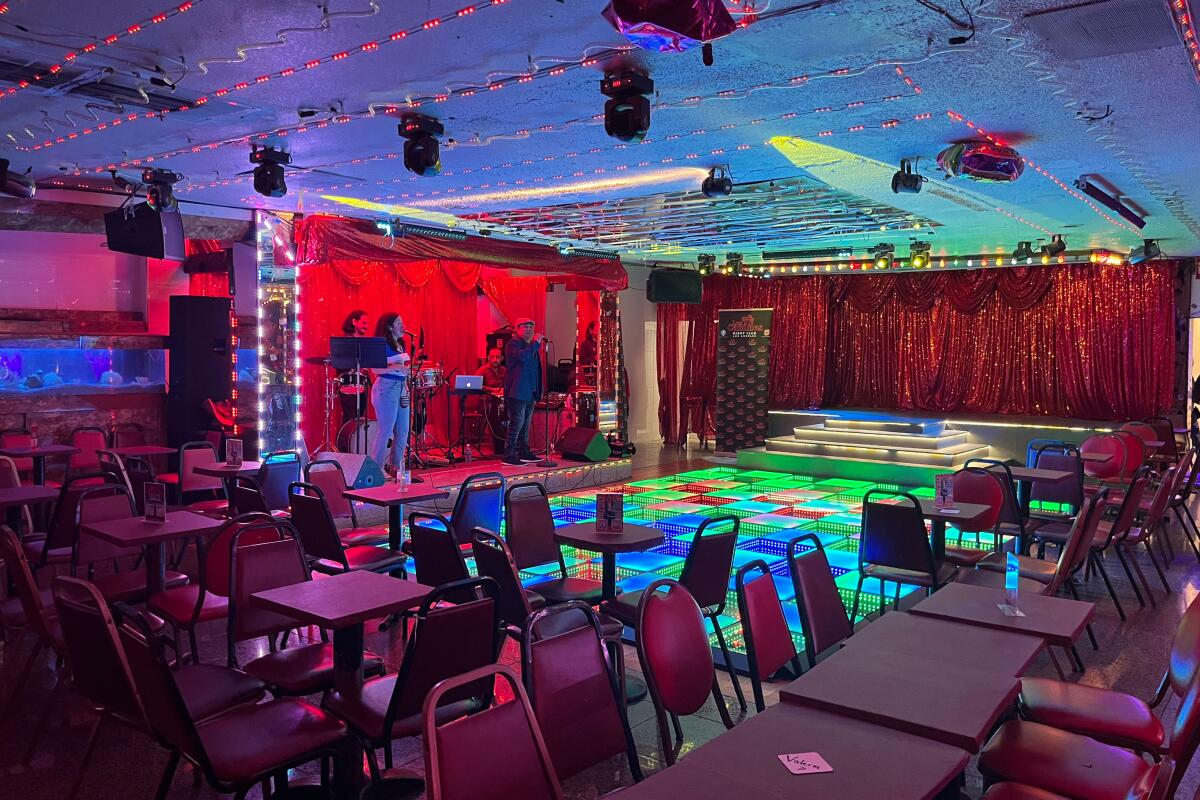 A colorfully lighted bar with tables and chairs, a dance floor and people performing onstage.