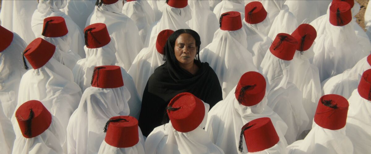 Islam Mubarak, dressed in black, in a sea of figures shrouded in white wearing red fezzes in "You Will Die at Twenty."