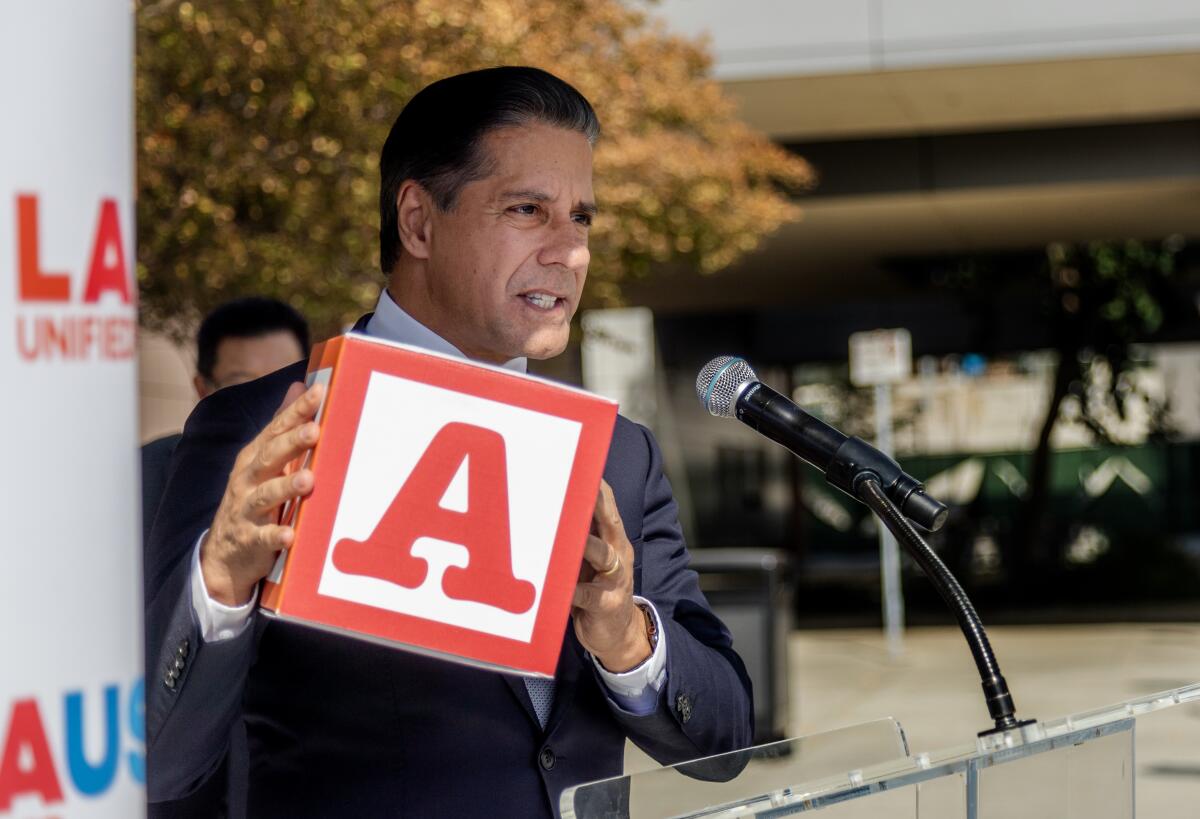 A man in a suit speaking into a microphone while holding up a red-and-white box resembling a child's wooden alphabet block