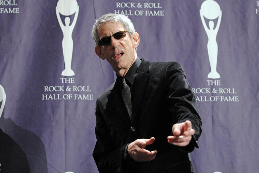 A man with gray hair wearing sunglasses and a black suit and pointing finger guns toward the camera