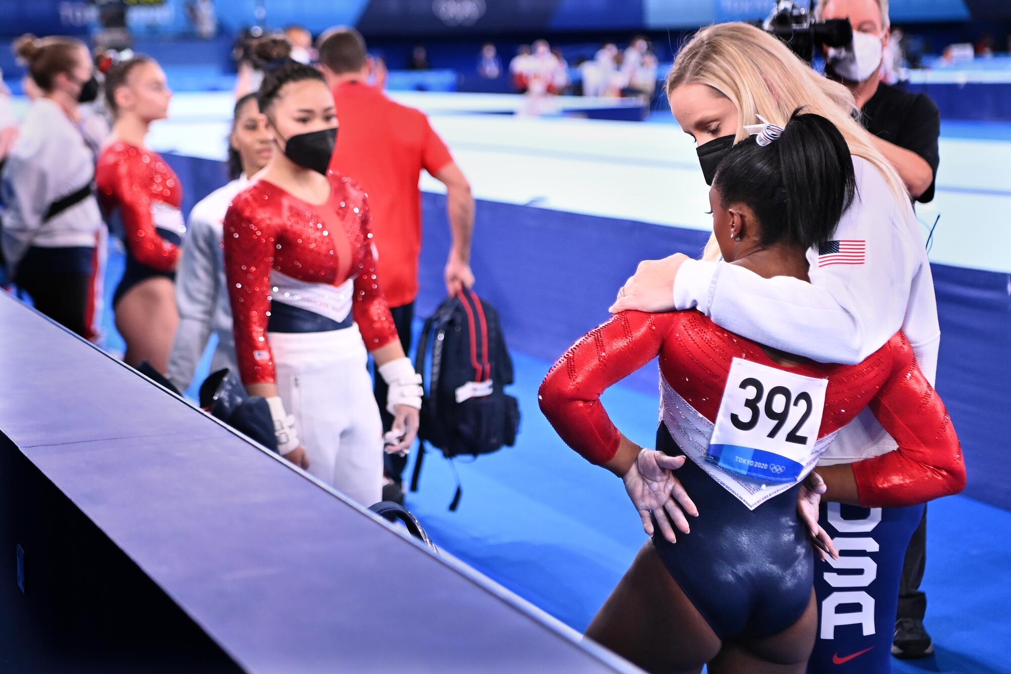 A woman in USA warmups puts her arm around Simone Biles, who stands with arms akimbo.