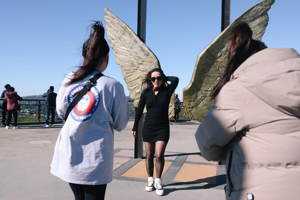 A woman posing in the wind as two people take her photo in front of a sculpture of angel wings