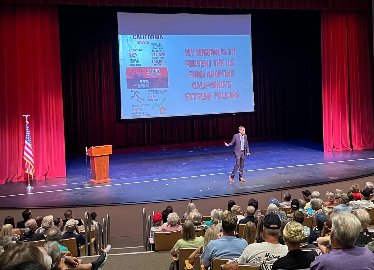 A man stands alone on a large stage, speaking to a packed audience. 