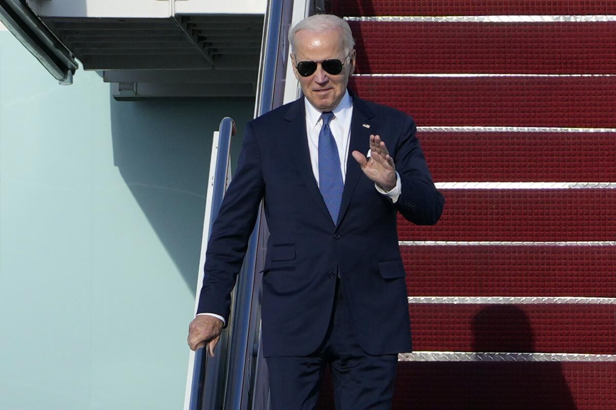 A man in sunglasses, dark suit and blue tie waves as he walks down steps