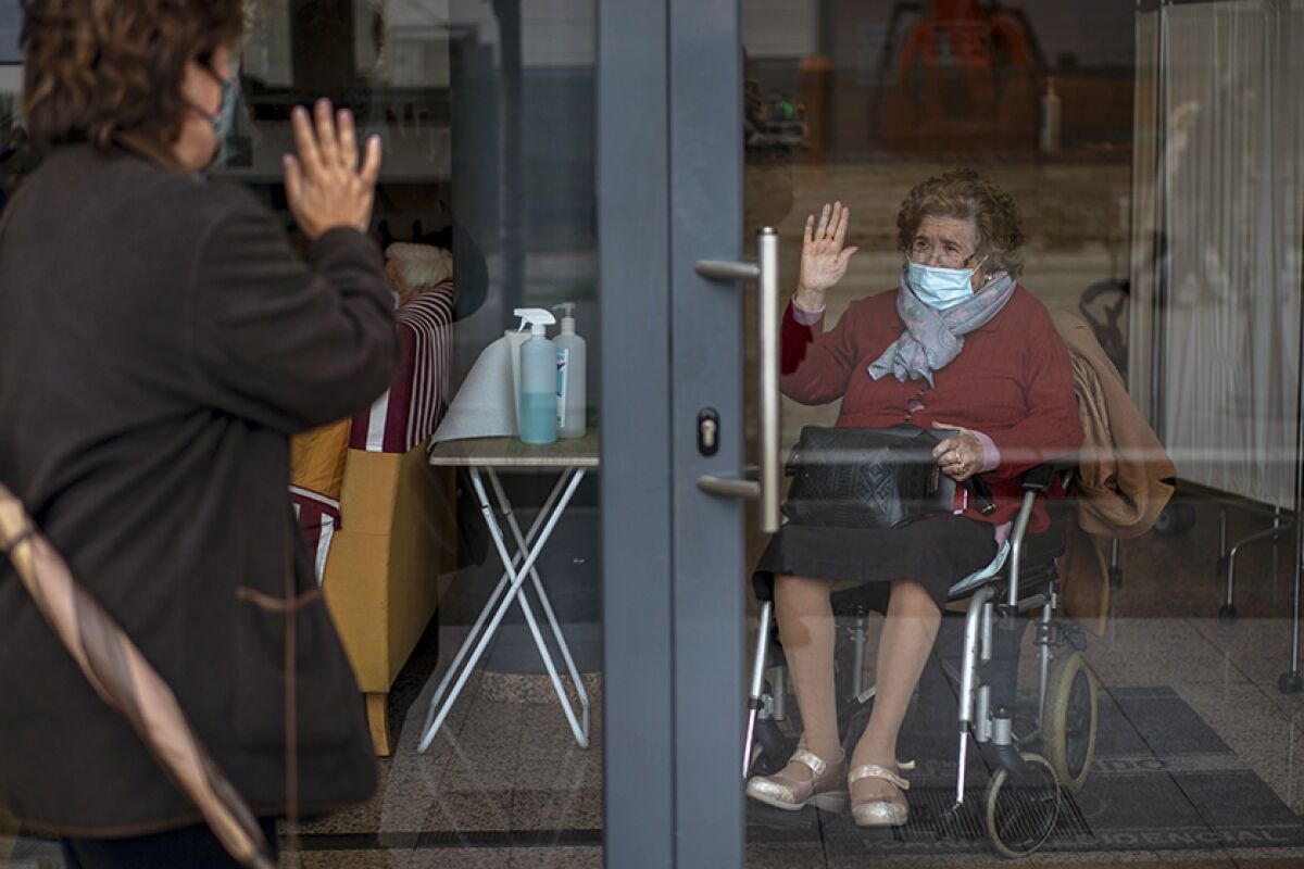 Masked woman waves goodbye to her masked mother, who is sitting in a wheelchair, through a glass door