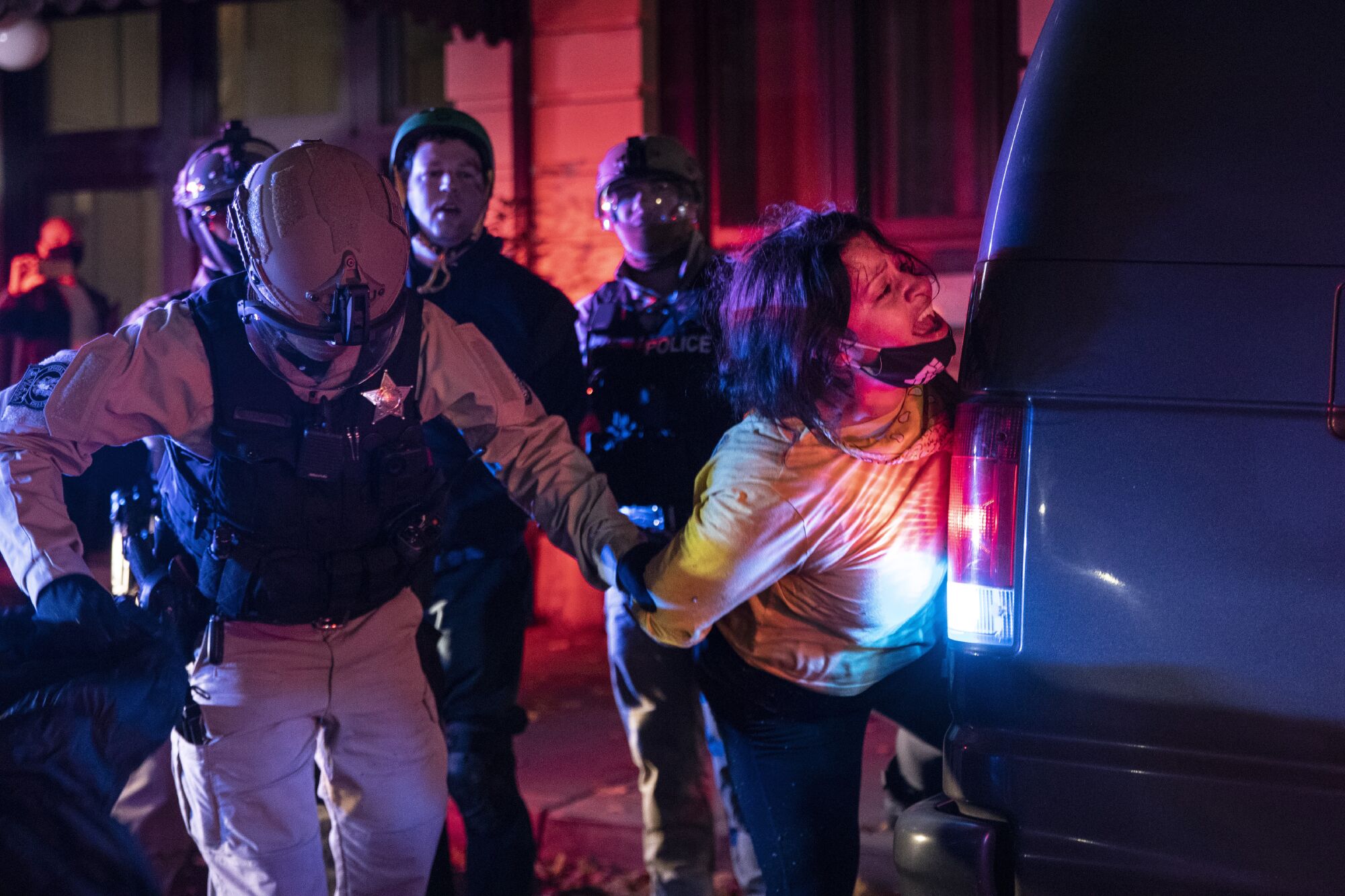 A woman with a mask on her chin has her hands pinned behind her back as she is pressed against a vehicle by police