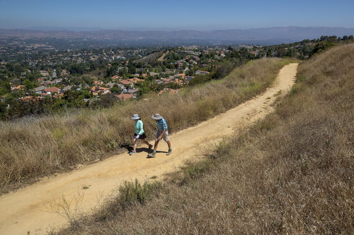 Two people walk on a trail between dry brush above a residential neighborhood