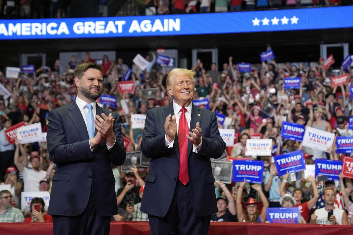 J.D. Vance in blue tie next to Donald Trump in red tie clapping before crowd