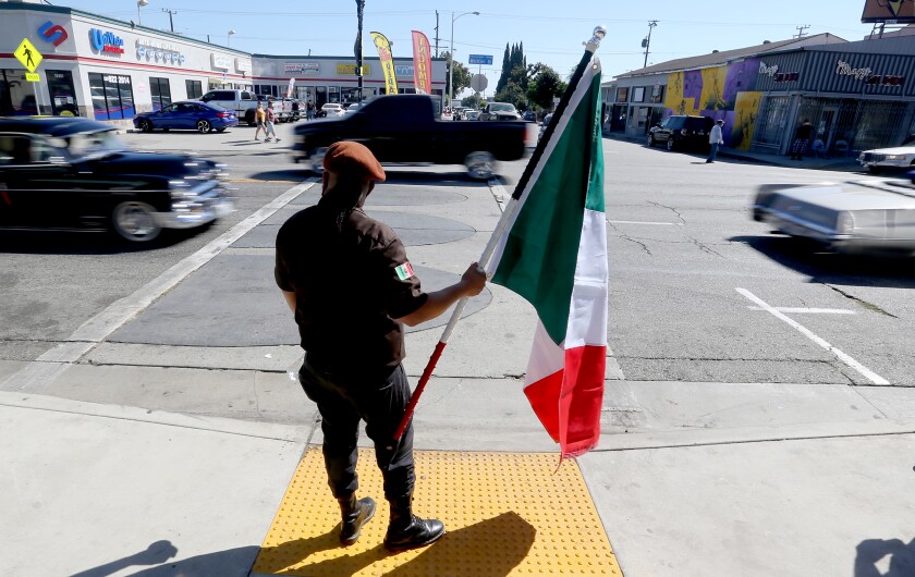 A man in the garb of the Brown Berets holds a Mexican flag on the sidewalk as cars pass on Whittier Boulevard