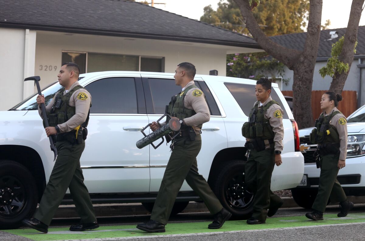 Deputies in flak vests carrying forced entry equipment walk in a street