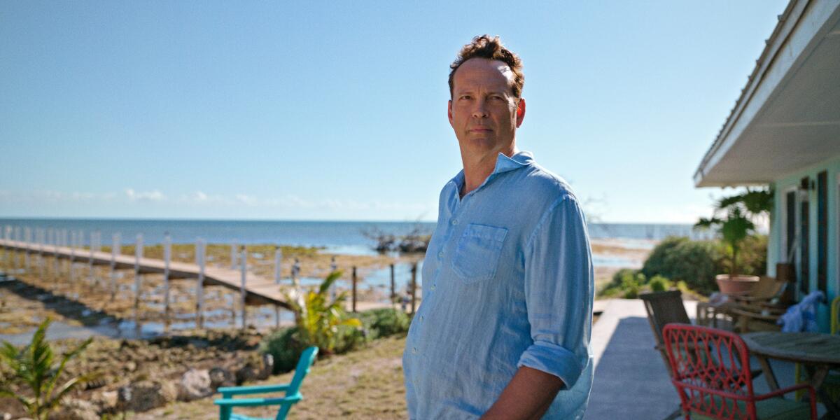 A man in a blue shirt standing on a back porch with the ocean behind him.