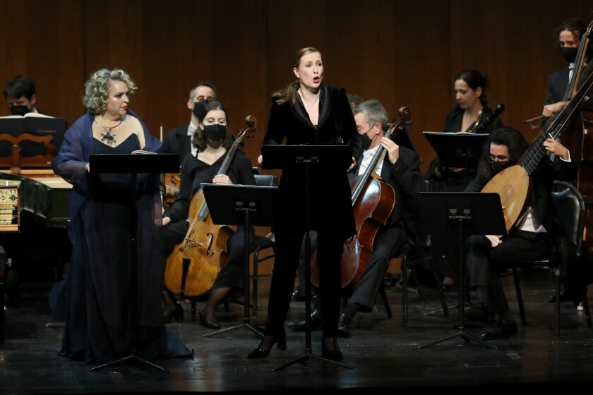Two women stand at music stands singing, with musicians playing instruments behind them. 