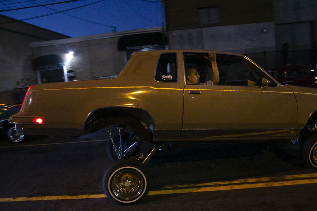 A man in the passenger window of a lowrider car with its rear hydraulics lifted a few feet off the street