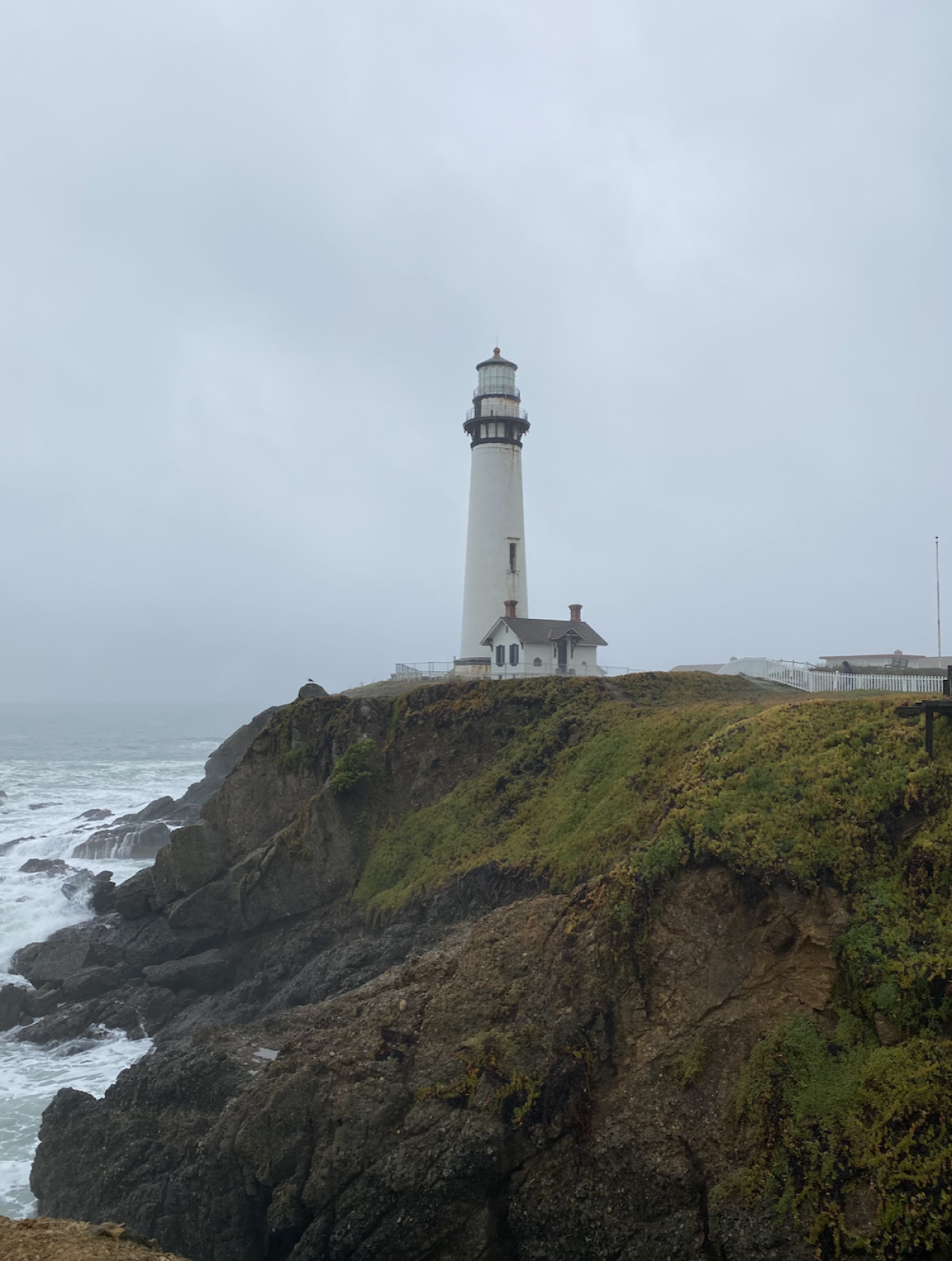 A lighthouse on the edge of an oceanside cliff under foggy skies.