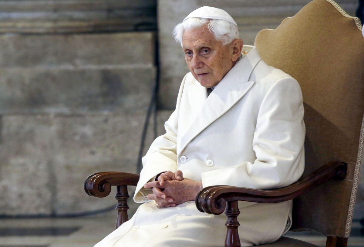 A white-haired man in a white cap and coat is seated with his hands clasped 