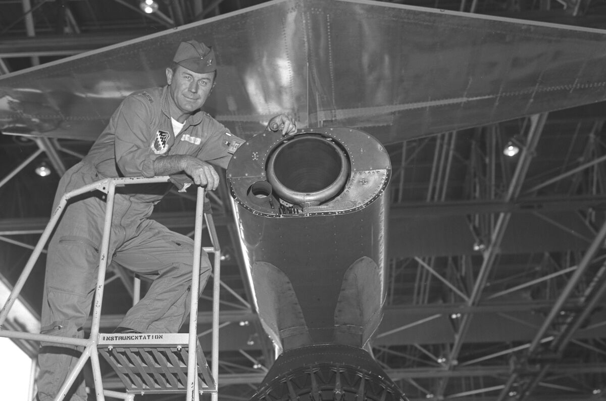A man in military cap and coveralls stands on a ladder, leaning an arm onto part of an aircraft.