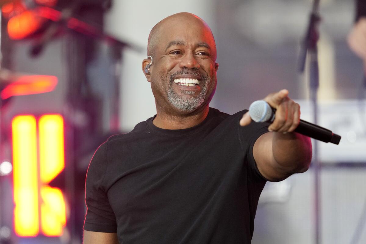 Darius Rucker in a black T-shirt smiling and pointing with a microphone in one hand