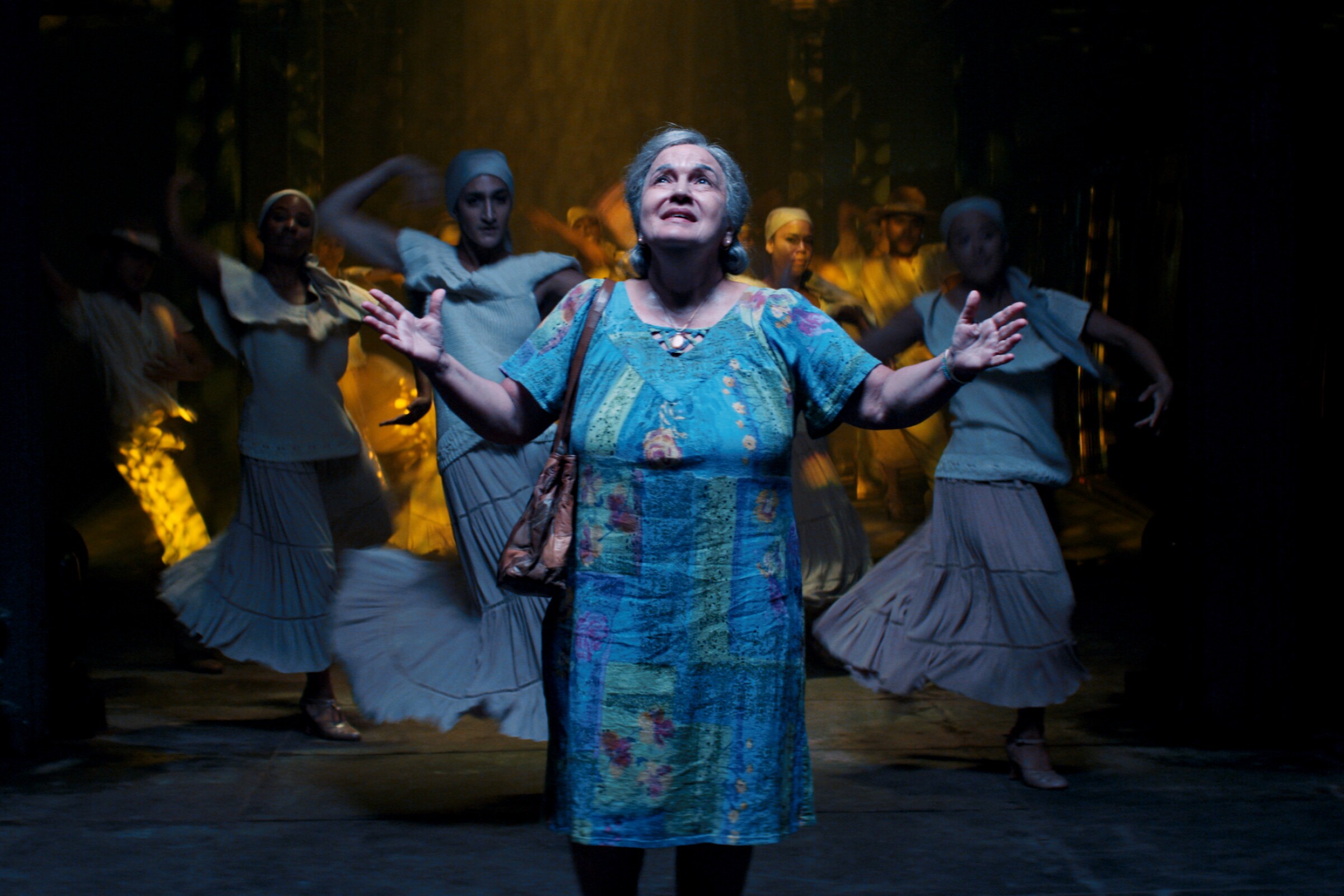 A woman in a housecoat with outstretched arms with dancers behind her