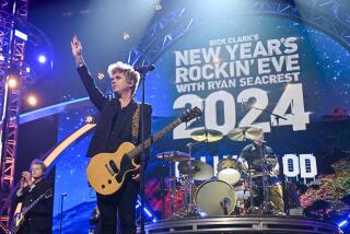 Billie Joe Armstrong holds his hand up with a guitar slung around his shoulder alongside Green Day bandmate Tré Cool