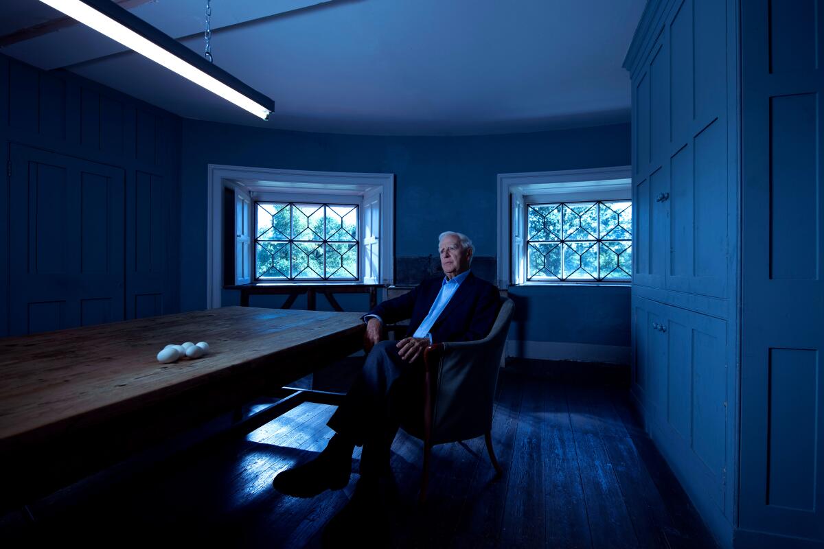 A man seated at a table is interviewed in a dimly lit room with two windows at its far end