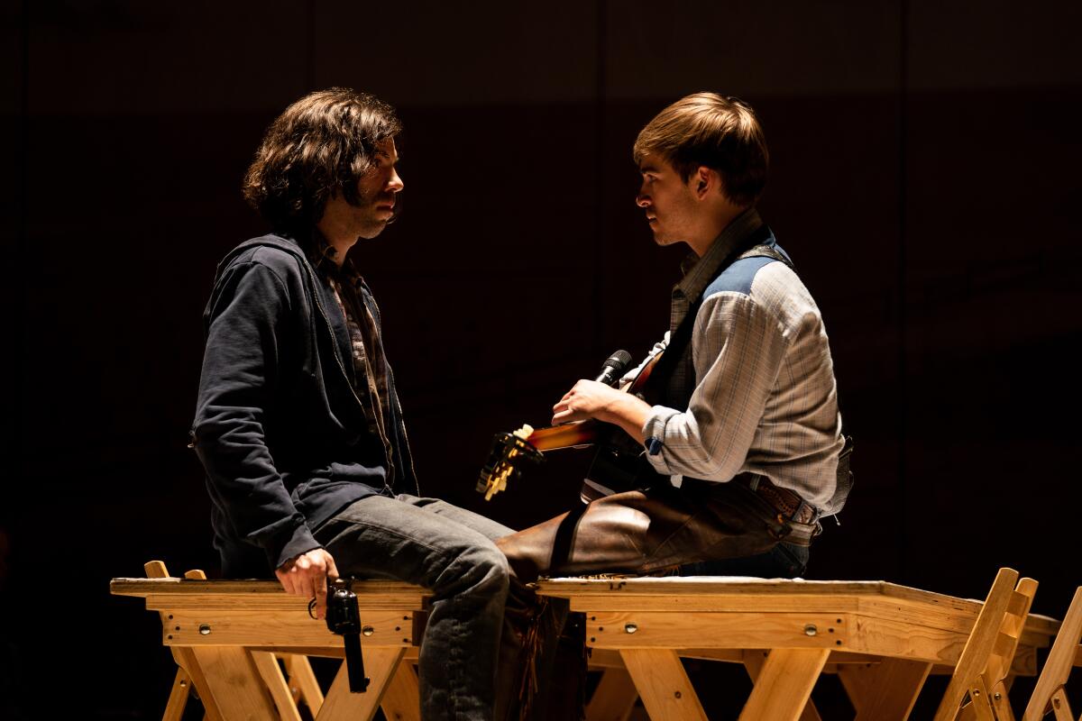 Two men, one holding a gun at his side, sit across from one another.