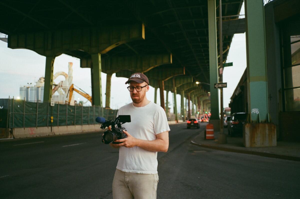 A man in a white T-shirt holding a camera under elevated train tracks