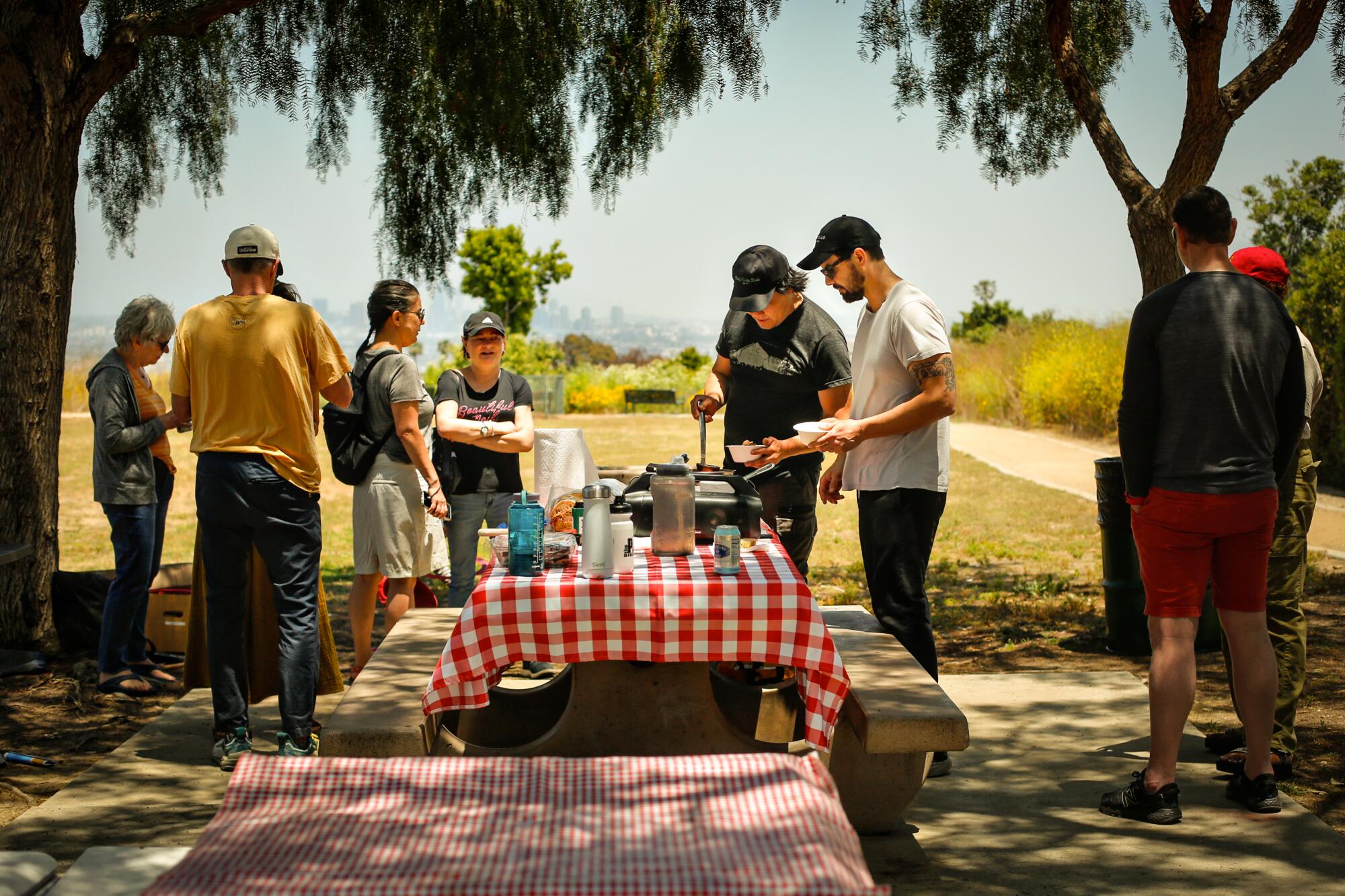 People mill about a picnic table covered with a red-checkered tablecloth.