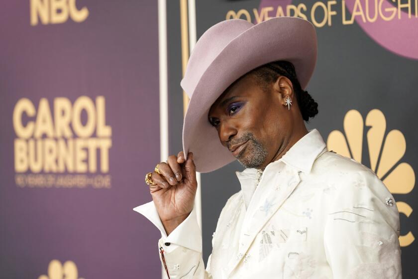 Billy Porter in a white suit tipping a dusty pink hat