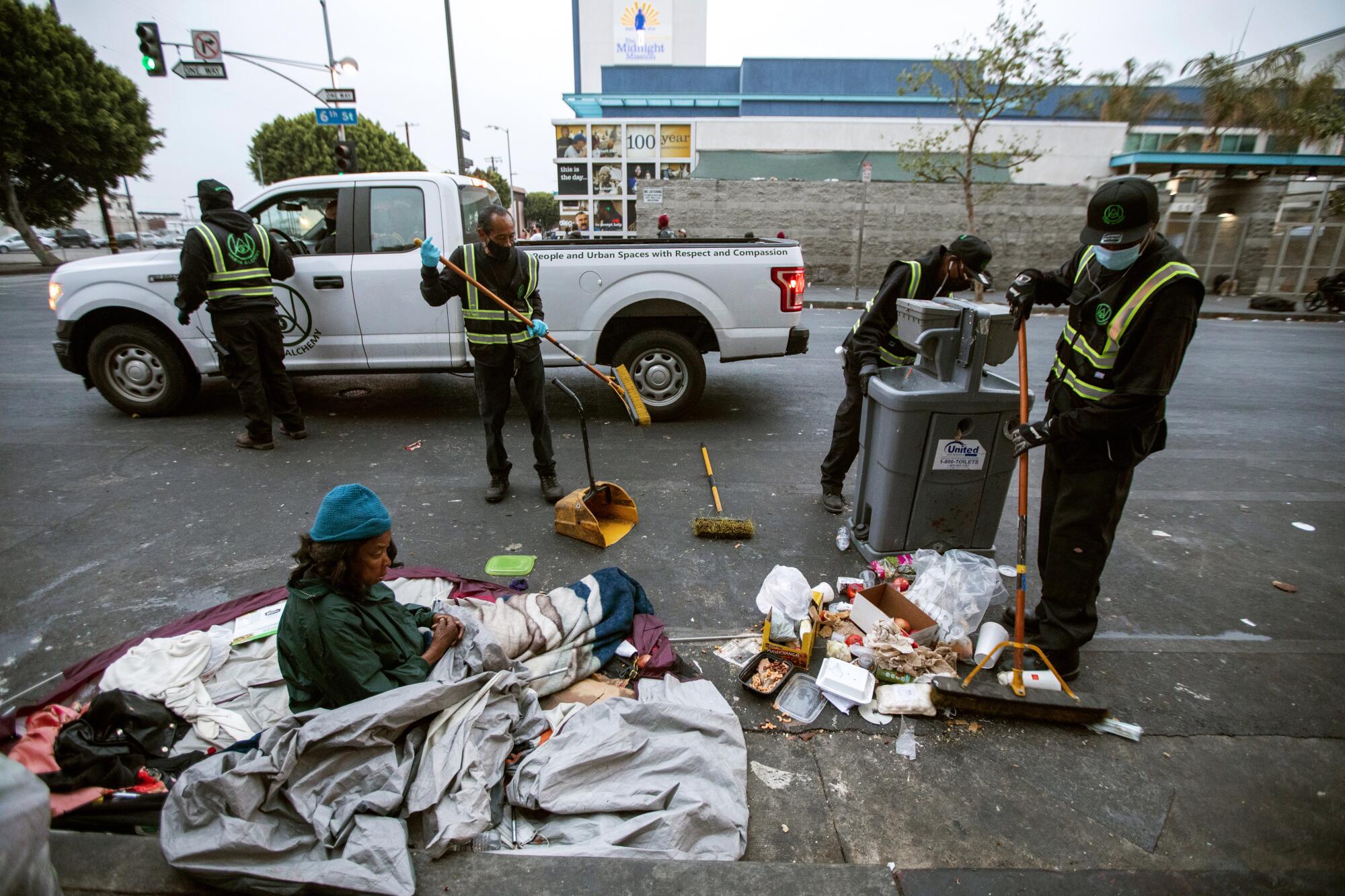 Workers carefully clean around a woman who is waking up on the sidewalk in the early morning in skid row