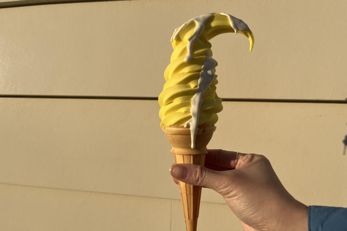 A hand holds a tall cone of lemon soft serve drizzled with white chocolate